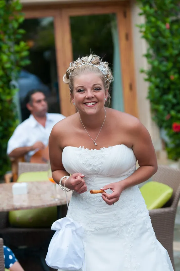 Smiling bride in a white strapless wedding dress holding a small snack outdoors.