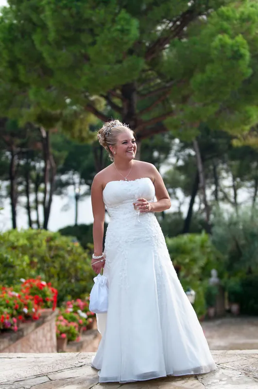 Smiling bride in strapless white wedding gown holding a glass and small white bag outdoors with green trees and flowers in background.