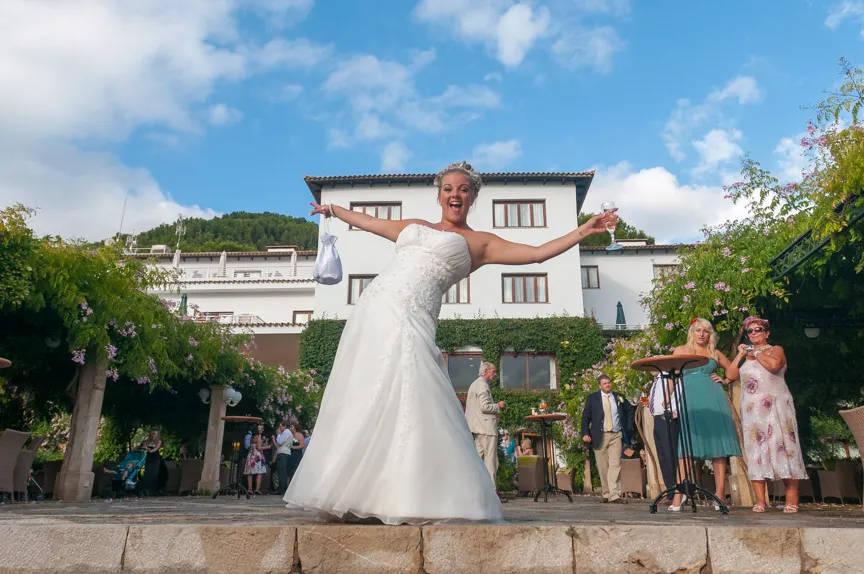 Bride in a white wedding dress smiling broadly with arms outstretched, holding a bouquet and a glass, outdoors in front of a building with guests in the background.