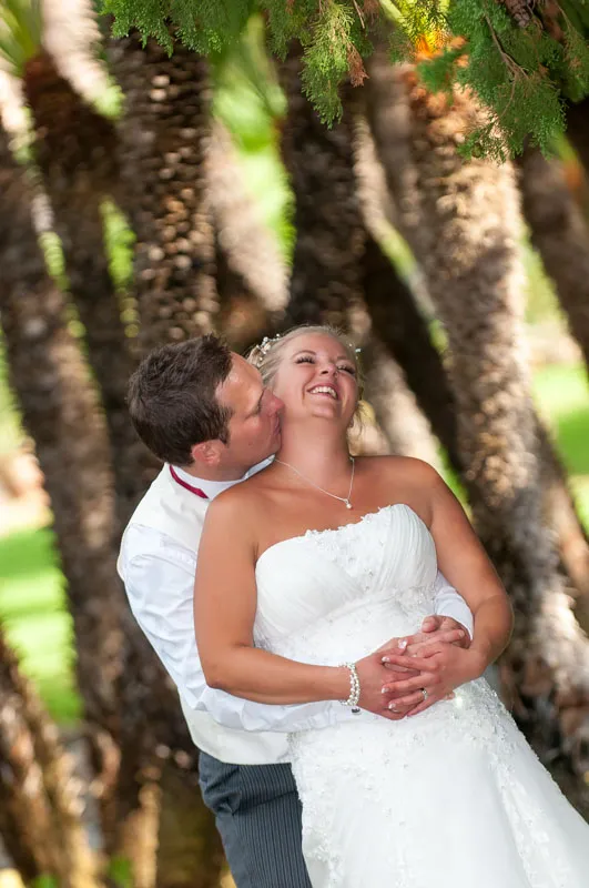 Groom in white shirt and vest embracing bride in strapless white wedding gown under palm trees, both smiling.