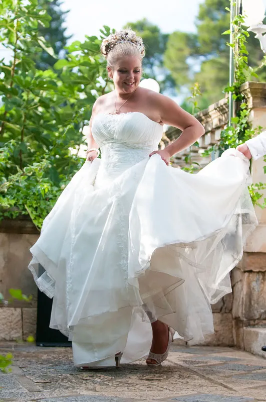 Smiling bride in a white strapless wedding gown holding up her dress outdoors with greenery and stone walls in the background.
