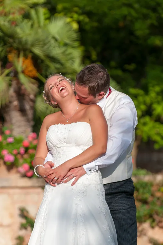 Bride laughing joyfully as groom hugs and kisses her from behind outdoors with greenery and flowers in the background.