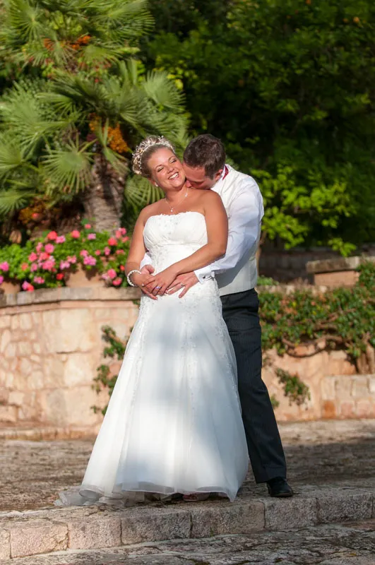 Bride in white wedding dress smiling while groom in white shirt embraces her from behind in an outdoor garden setting.