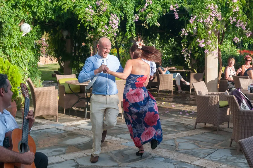 A man in a light blue shirt and beige pants dances outdoors with a woman in a blue dress with large pink flowers under a flowering green canopy.