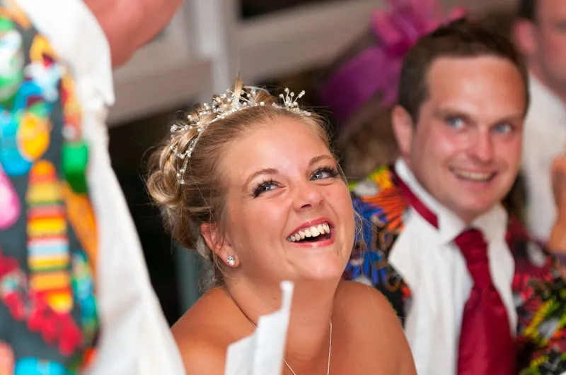 Smiling bride wearing a tiara and earrings at a wedding reception, with a man in a colorful jacket and tie in the background.