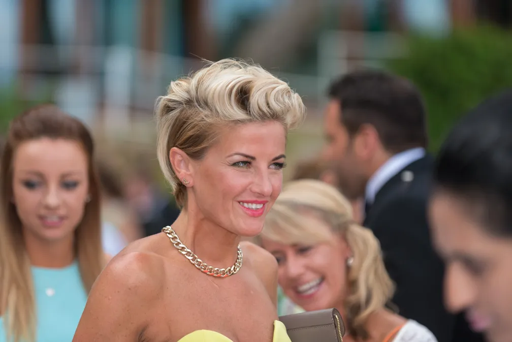 Smiling woman with short blonde hair and a gold chain necklace at a social event with blurred people in the background.