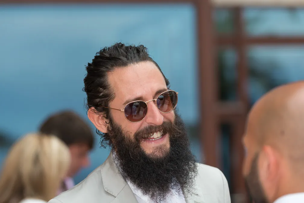 Man with dark beard, moustache, and sunglasses smiling in conversation at an outdoor event.