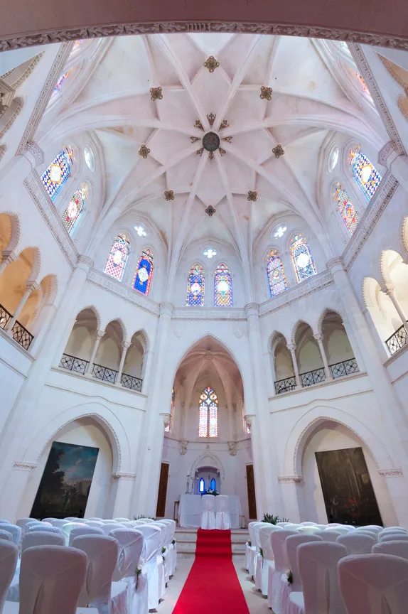 Interior of a bright chapel with a red carpet aisle leading to an altar, surrounded by white chairs and tall stained glass windows.