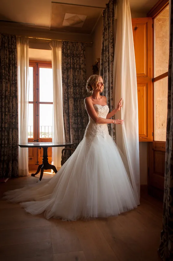 Bride in a strapless white wedding gown standing by a window with curtains, smiling and looking outside in a warmly lit room.