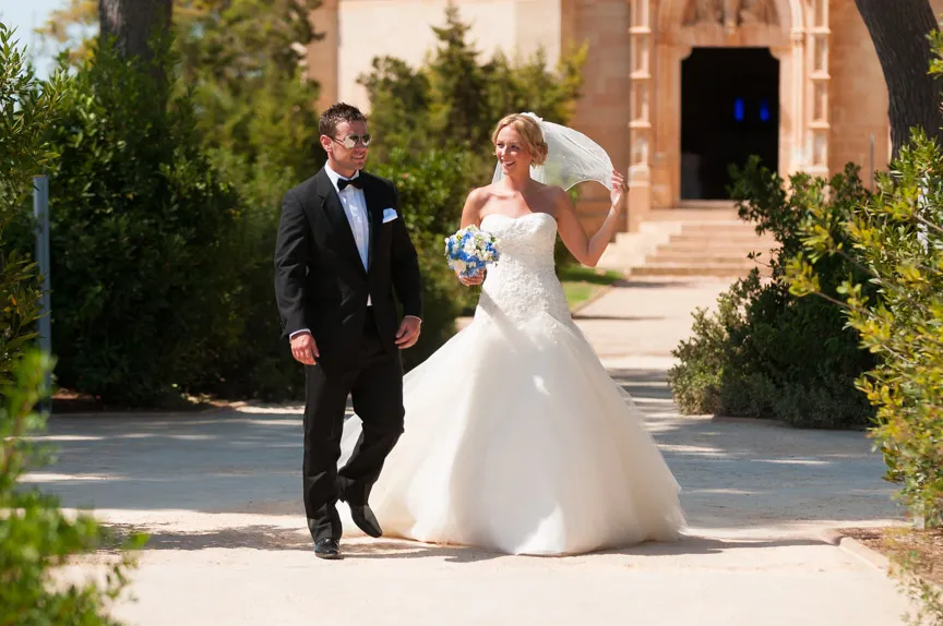 Bride in white wedding dress holding a bouquet and veil walking beside groom in black tuxedo outdoors near a church entrance.