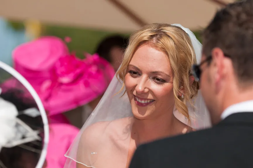 Close-up of a smiling bride in a veil looking at the groom during an outdoor wedding ceremony with a bright pink hat visible in the background.