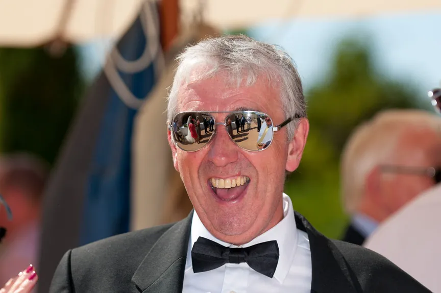 Smiling older man wearing a tuxedo and reflective sunglasses at an outdoor event.
