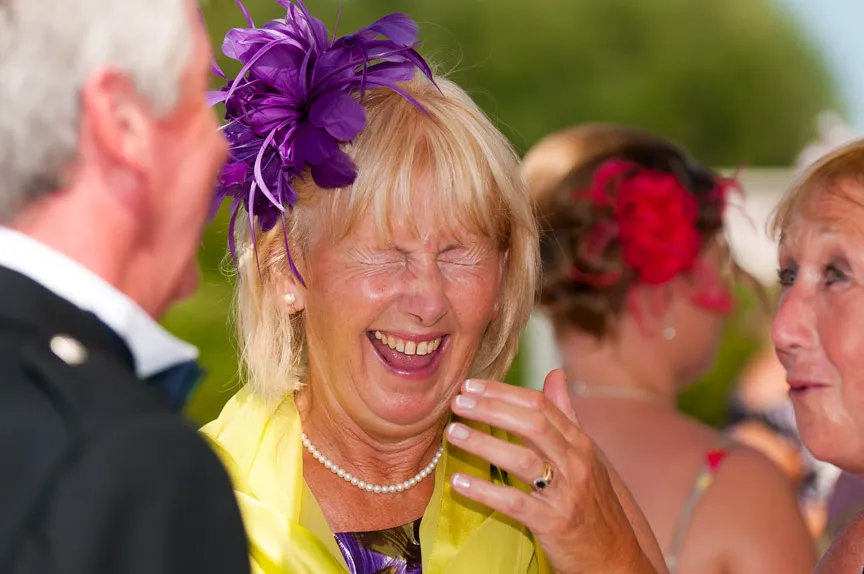 Older woman laughing with eyes closed, wearing a purple feathered headpiece and yellow scarf, talking with others.