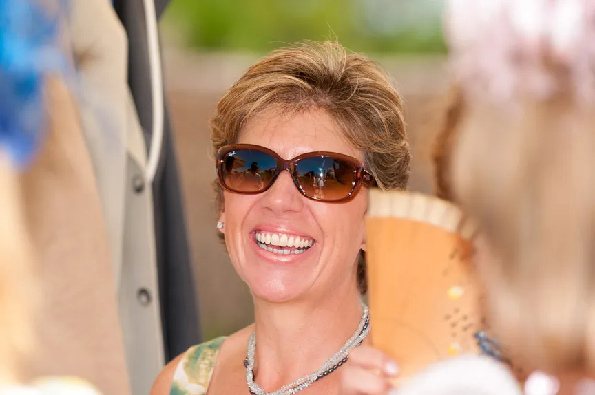 Woman wearing sunglasses smiling outdoors with blurred background and another person holding a wooden fan in the foreground.