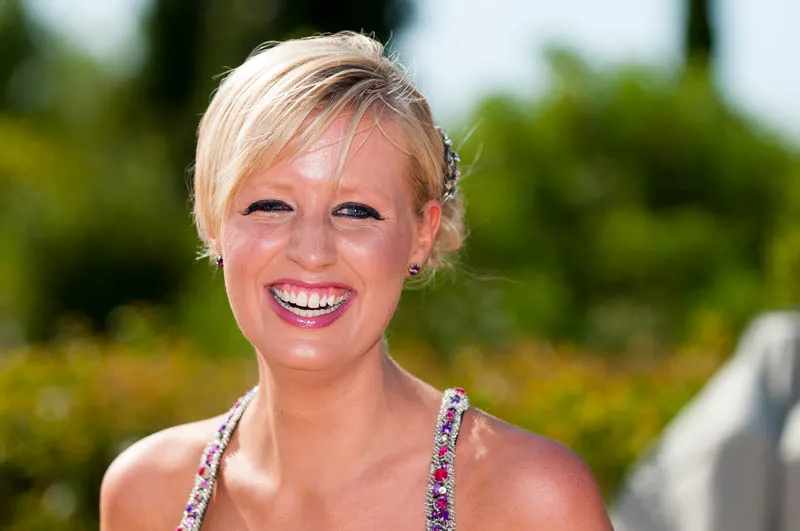 Smiling blonde woman outdoors with a jeweled strap dress and blurred greenery background.