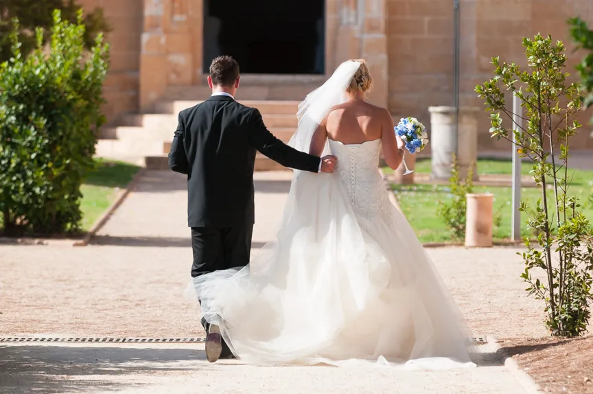 Bride in a white wedding gown holding a bouquet and groom in a black suit walking away toward a building entrance.