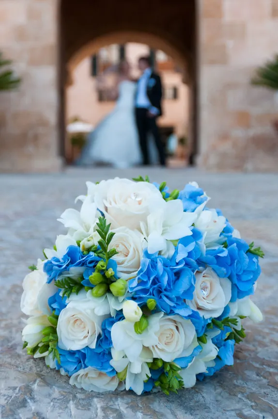 Close-up of a bridal bouquet with white roses and blue flowers on a stone surface, with a blurred bride and groom in the background.