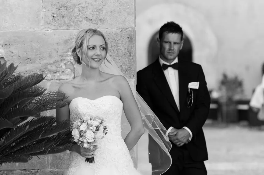 Black and white photo of a bride holding a bouquet and smiling, standing near a stone wall, with a groom in a tuxedo blurred in the background.