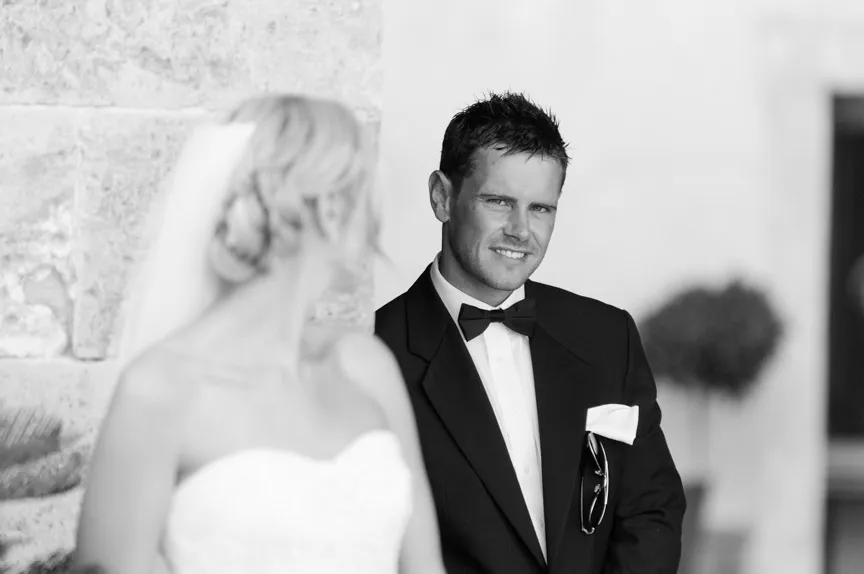 Black and white photo of a groom in a tuxedo smiling at a bride in a wedding dress with a veil.