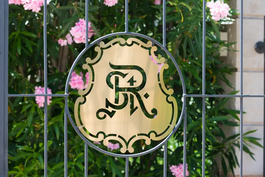 Decorative metal gate with a circular golden emblem featuring stylized letters and the number 24, with green foliage and pink flowers in the background.