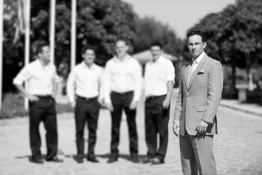 Man in a suit stands sharply in focus in front of four men in white shirts and dark pants who are blurred in the background on a paved outdoor path with trees.
