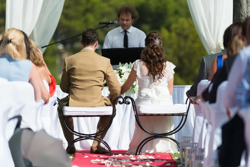 Bride and groom holding hands seated during outdoor wedding ceremony with officiant speaking at altar.