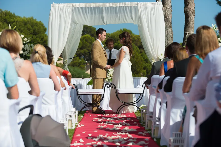 Bride and groom holding hands under a white wedding arch during outdoor ceremony with guests seated on either side.
