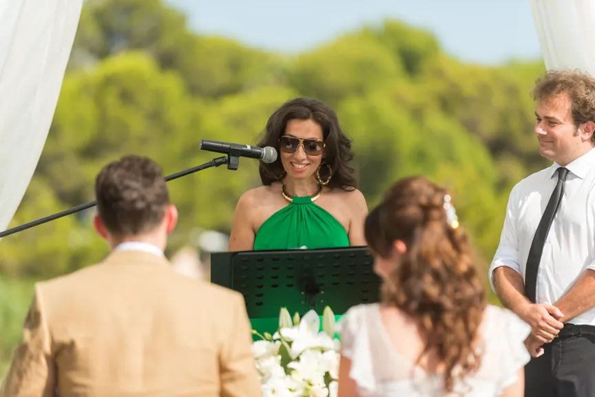 Woman in green dress and sunglasses speaking at a microphone during an outdoor wedding ceremony with the bride, groom, and officiant visible.