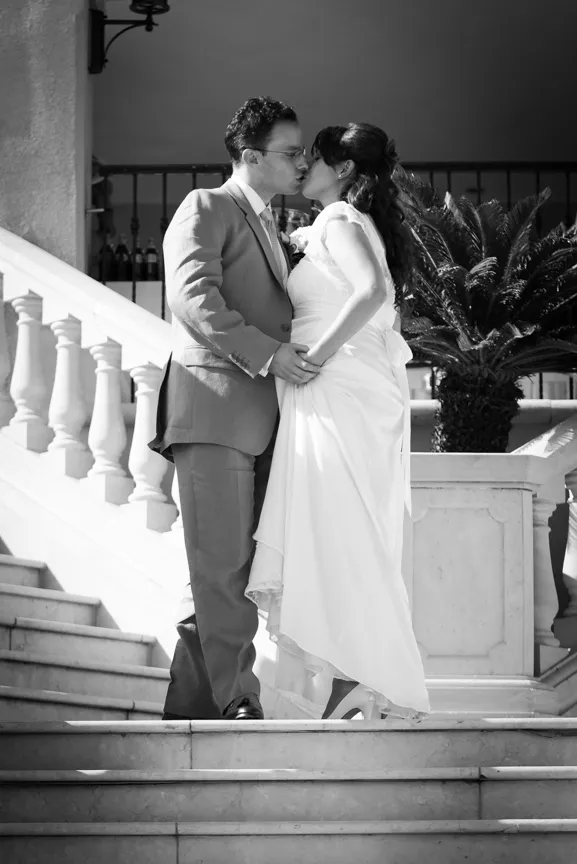 Bride and groom kissing on marble stairs, bride lifting her dress slightly, black and white photo.