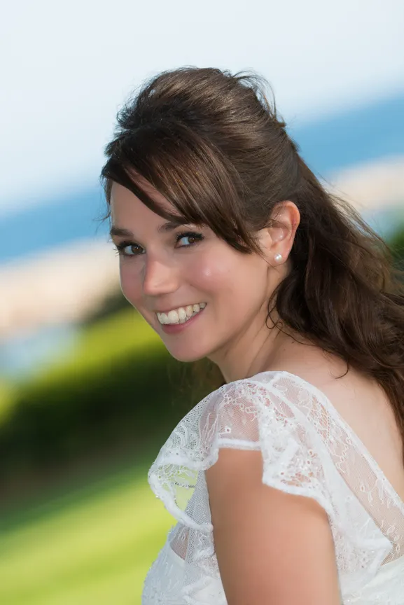 Smiling woman with brown hair in a white lace dress outdoors with a blurred green and blue background.