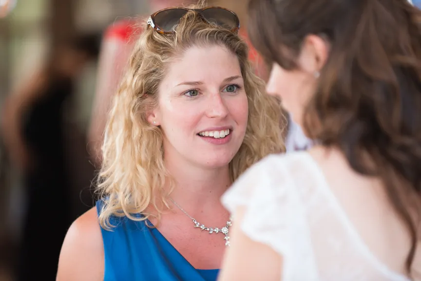 Smiling blonde woman with sunglasses on her head wearing a blue top and a silver necklace talking to a brunette woman in a white lace dress.