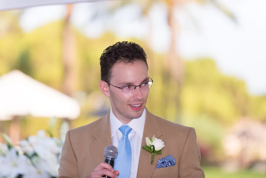 Man in tan suit jacket with blue tie and glasses holding a microphone, speaking outdoors with blurred greenery in the background.