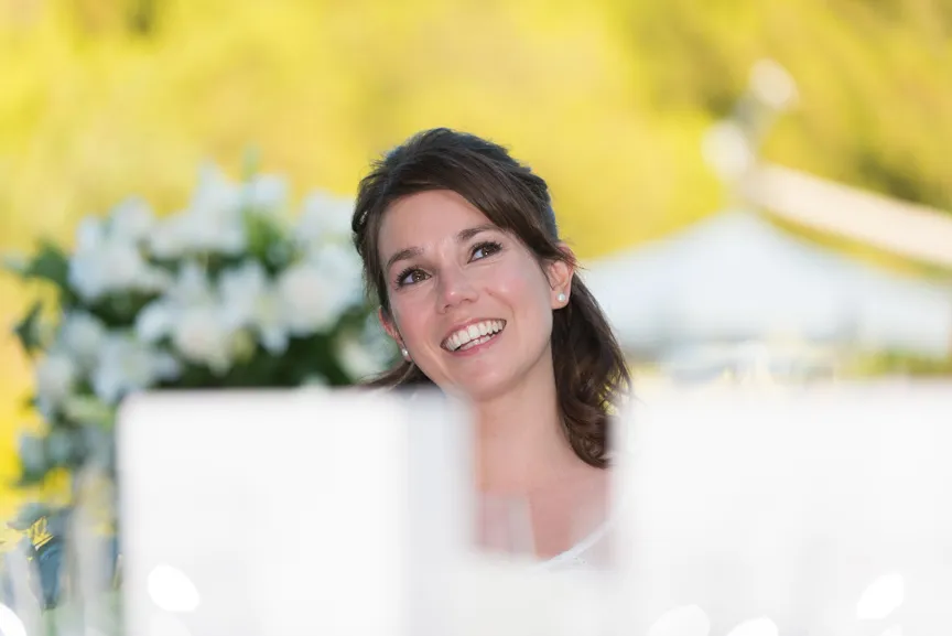 Smiling bride with dark hair and pearl earrings at a wedding reception table with white floral arrangements in soft background.