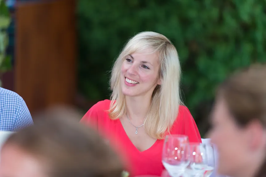 Smiling blonde woman in a red top seated at a table with wine glasses, blurred people in foreground.