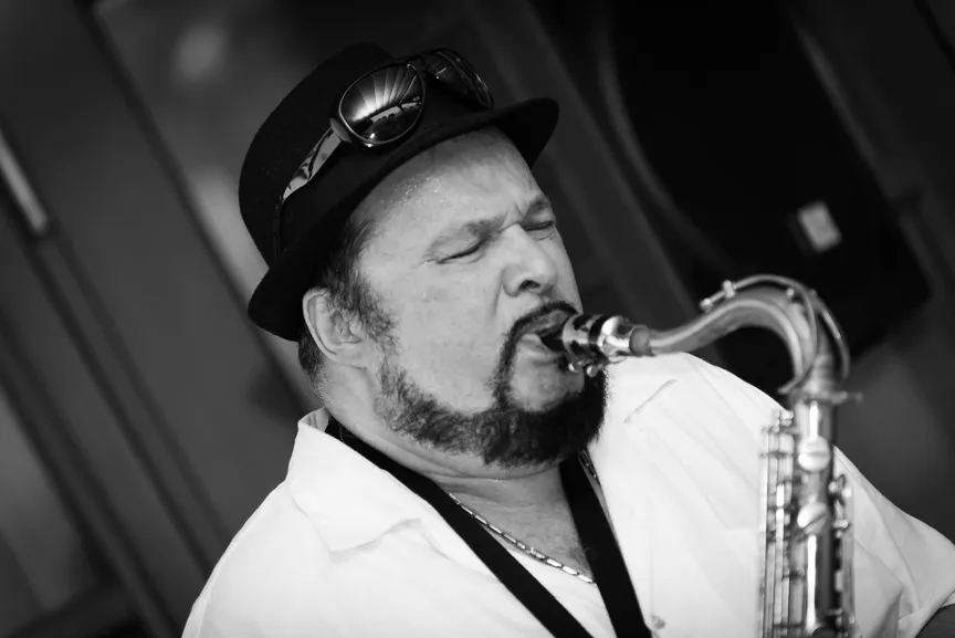 Black and white close-up of a man wearing a hat and sunglasses playing a saxophone with eyes closed.