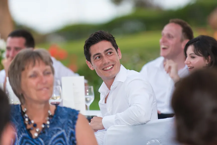 Group of people smiling and enjoying a social event, with a man in a white shirt prominently looking to the side.