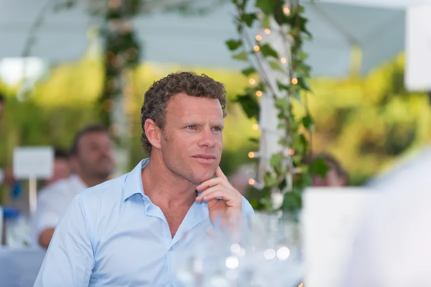 Man with curly hair wearing a light blue shirt, sitting thoughtfully at an outdoor event decorated with ivy and string lights.