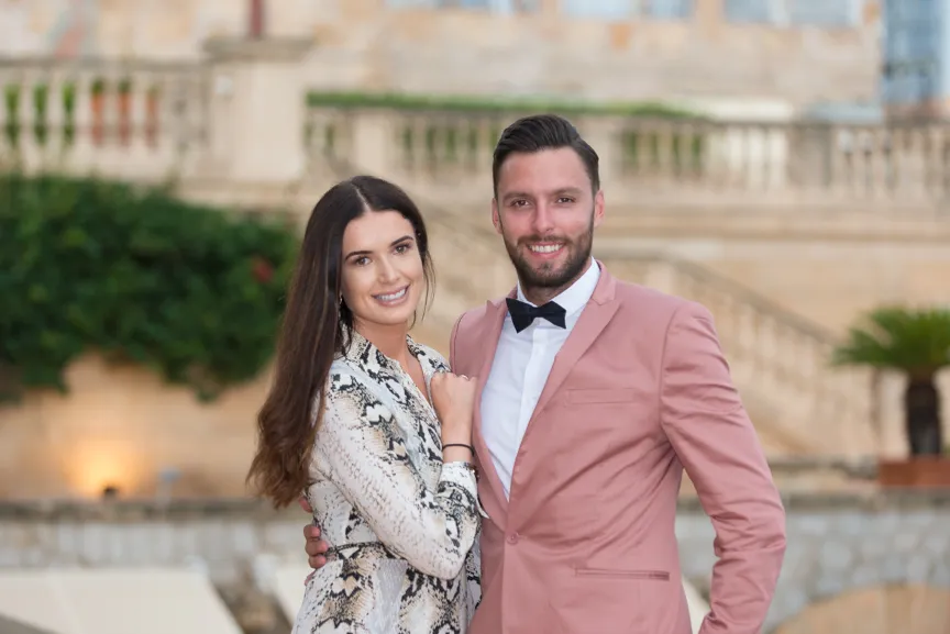 Smiling couple posing together outdoors, woman in patterned dress and man in pink suit with bow tie.