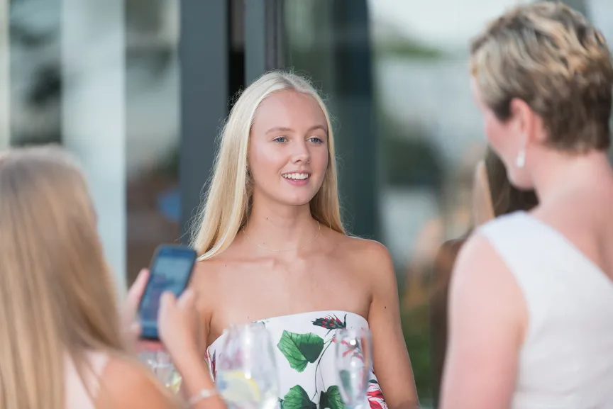Young woman with long blonde hair and floral off-shoulder dress smiling while facing two other women, one holding a smartphone.