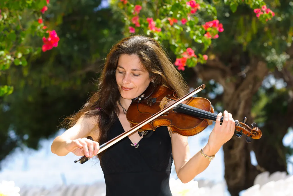 Woman in a black dress playing violin outdoors with pink flowers and trees in the background.