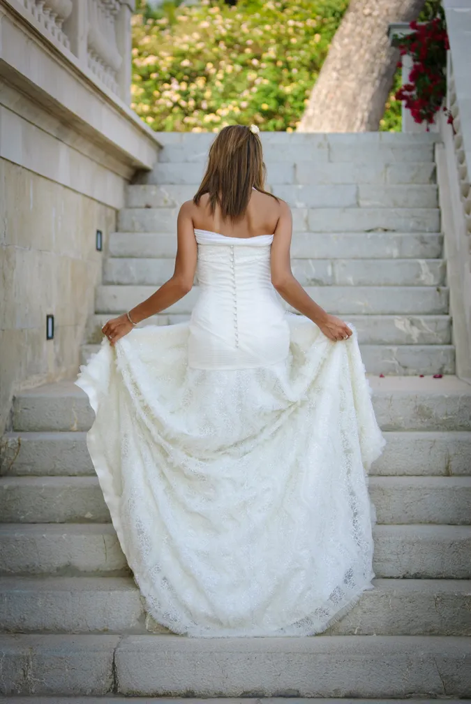 Bride in a strapless white wedding gown holding the train while walking up stone steps outdoors.