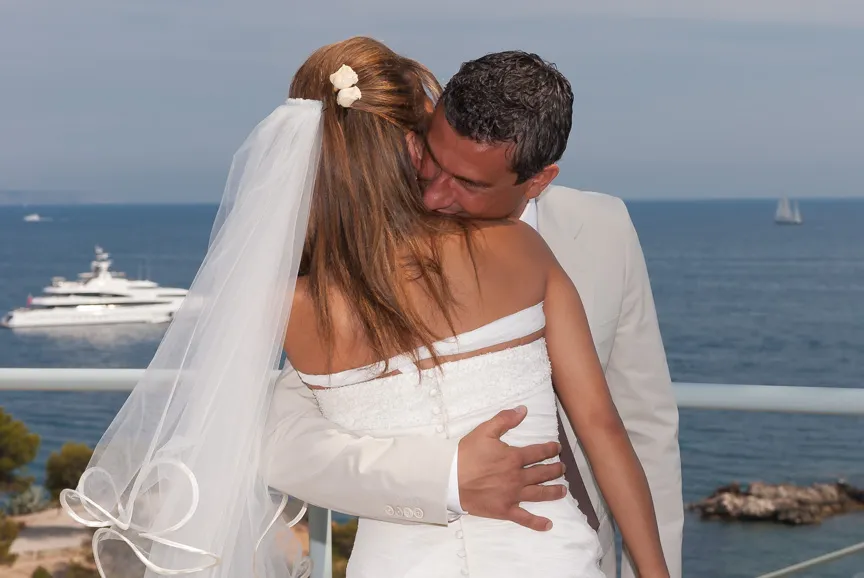 Bride and groom embracing on a balcony overlooking the ocean with a yacht in the background.