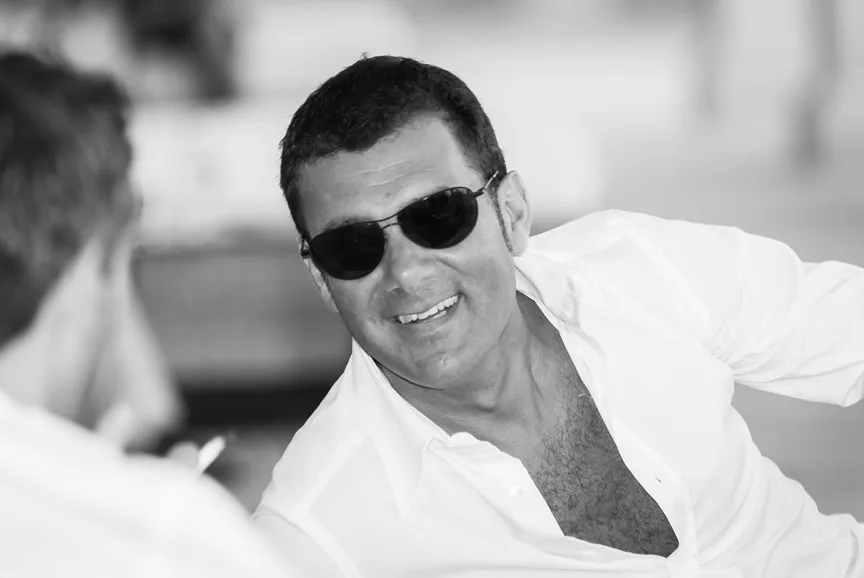 Smiling man wearing sunglasses and an open white shirt, leaning forward during a conversation.