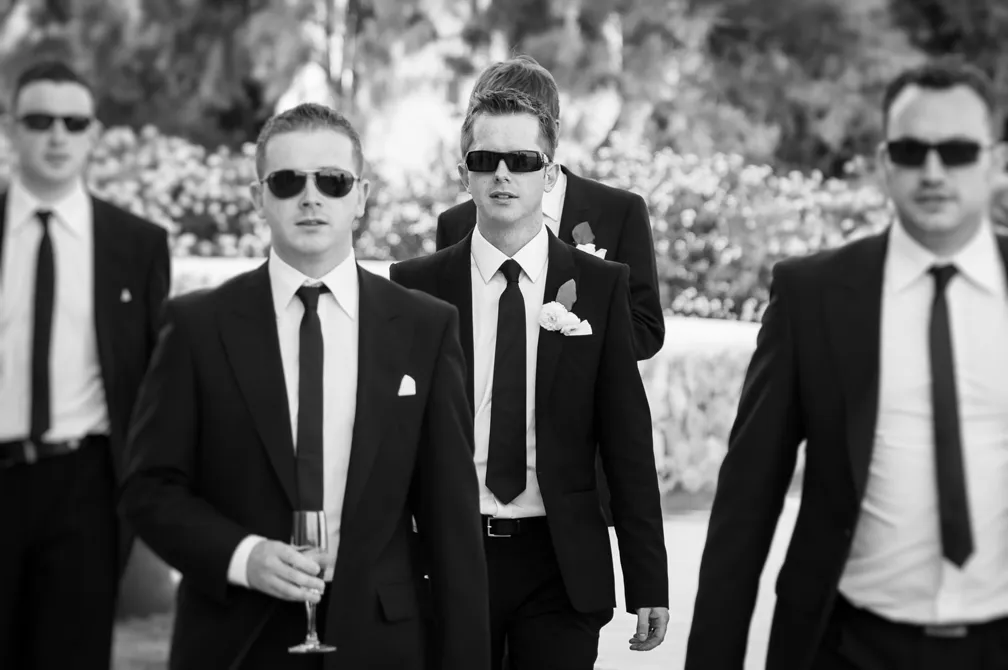 Group of men dressed in black suits and ties wearing sunglasses, walking outdoors, with one holding a glass of champagne.