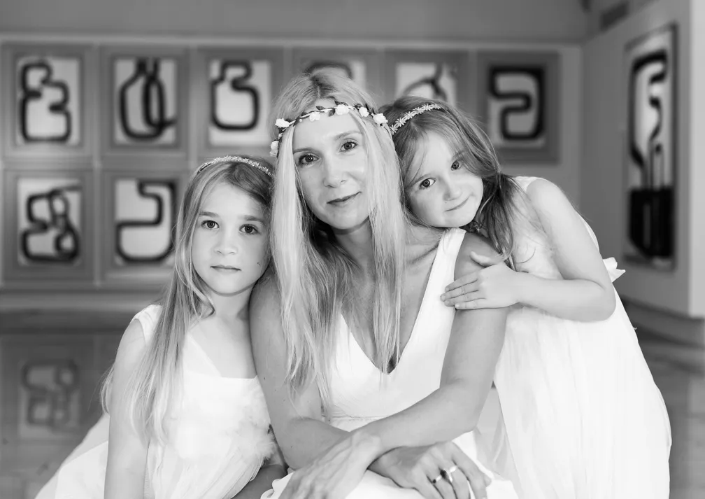 Black and white photo of a woman with long hair and two young girls in white dresses, all wearing floral headbands, posing indoors with abstract art in the background.