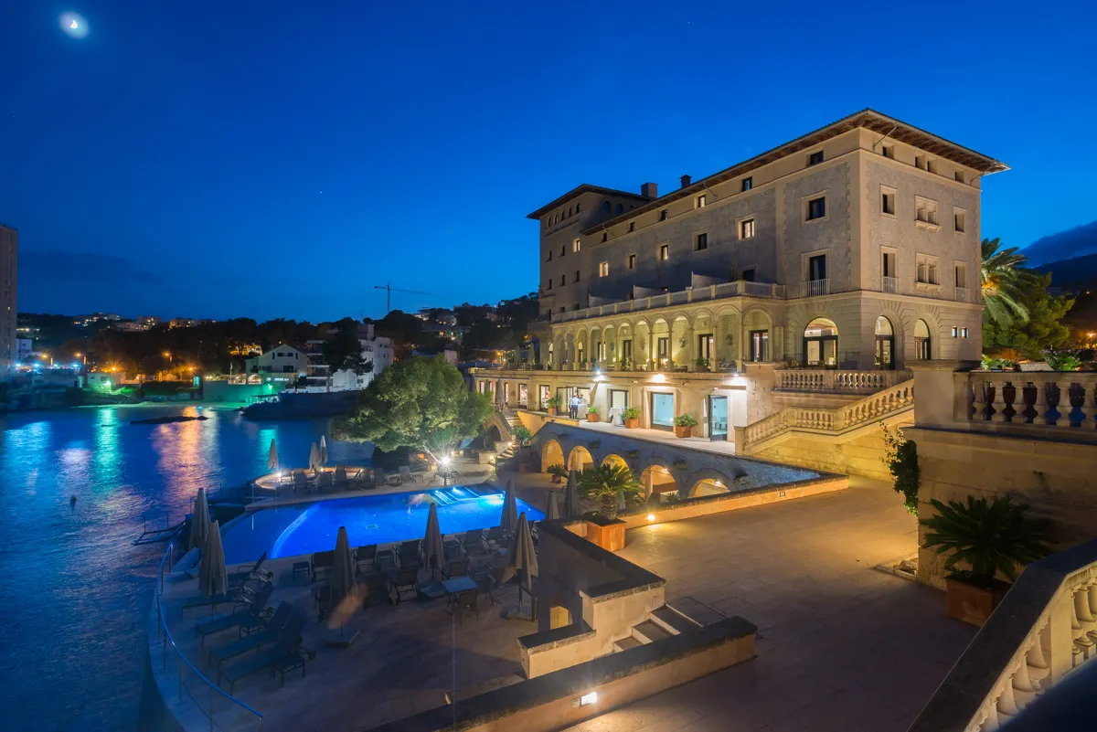 Elegant hotel building illuminated at dusk with a lit swimming pool and calm waterfront nearby under a crescent moon.