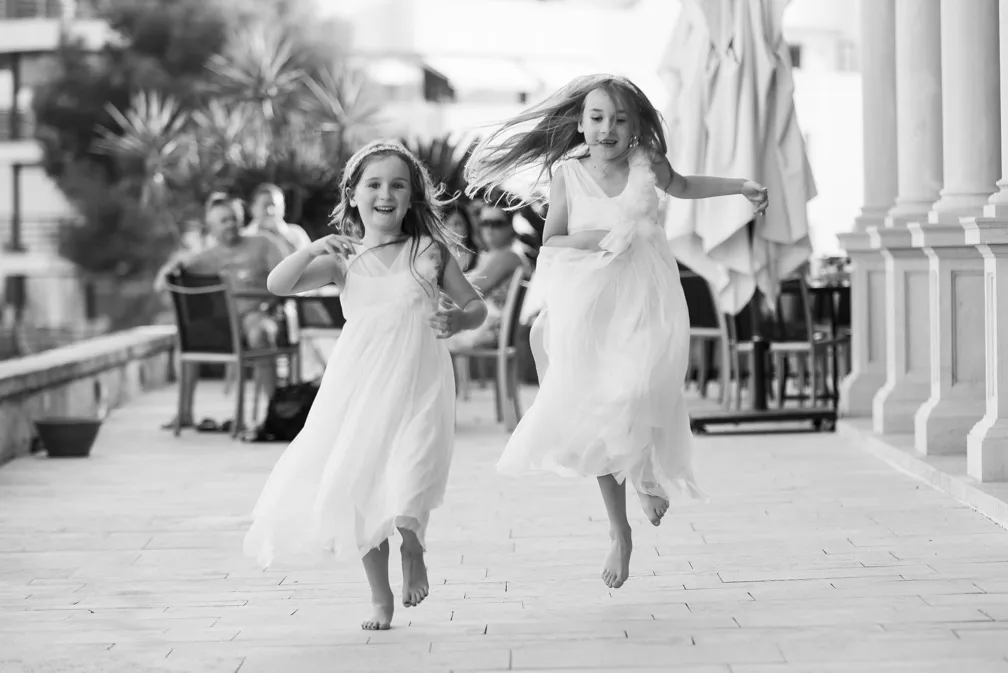 Two young girls in white dresses joyfully running barefoot on a paved outdoor walkway near seated people and columns.