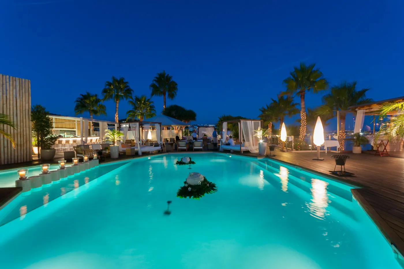 Illuminated outdoor pool at a luxury lounge with palm trees and cabanas under a deep blue evening sky.