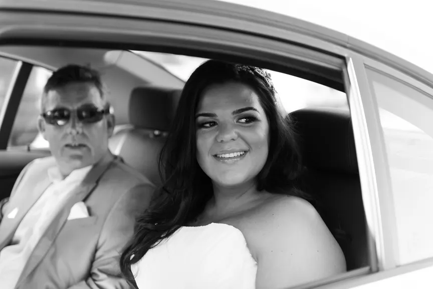 Smiling bride in a strapless wedding dress sitting in a car next to a man wearing sunglasses and a suit.