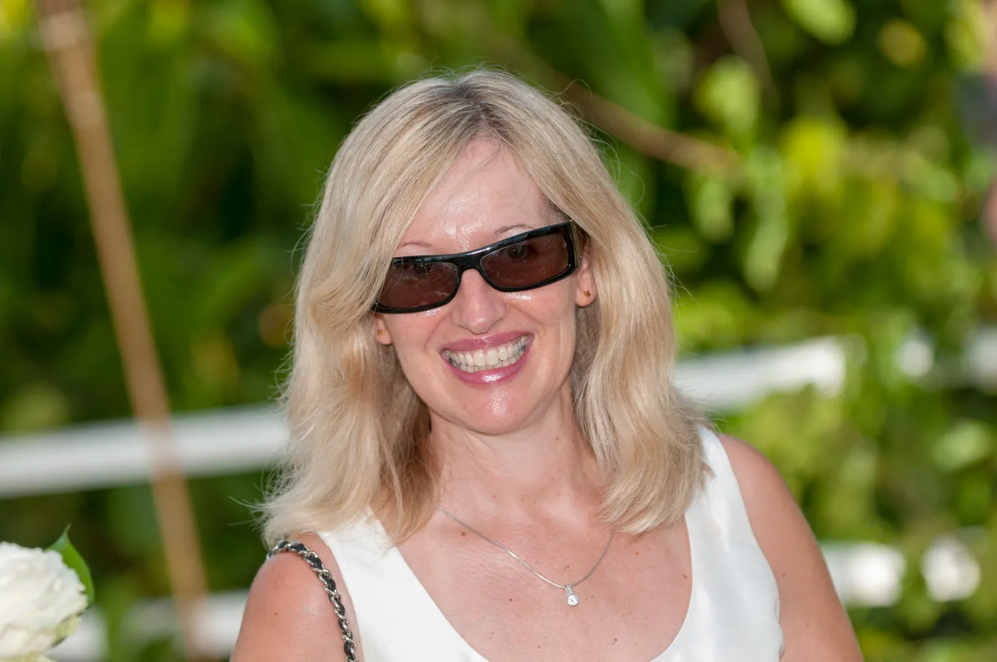 Smiling woman with blonde hair wearing dark sunglasses and a white sleeveless top outdoors.